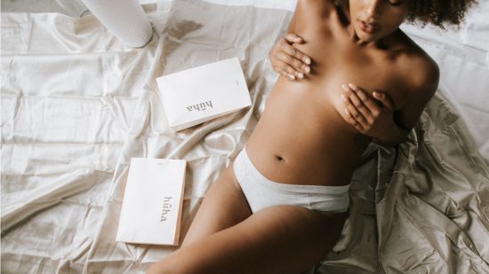 woman lying in bed topless, covering her chest, with gift boxes next to her.