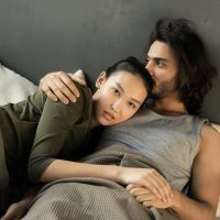 dark hair man holding asian woman intimately in grey bed talking about jackhammer sex