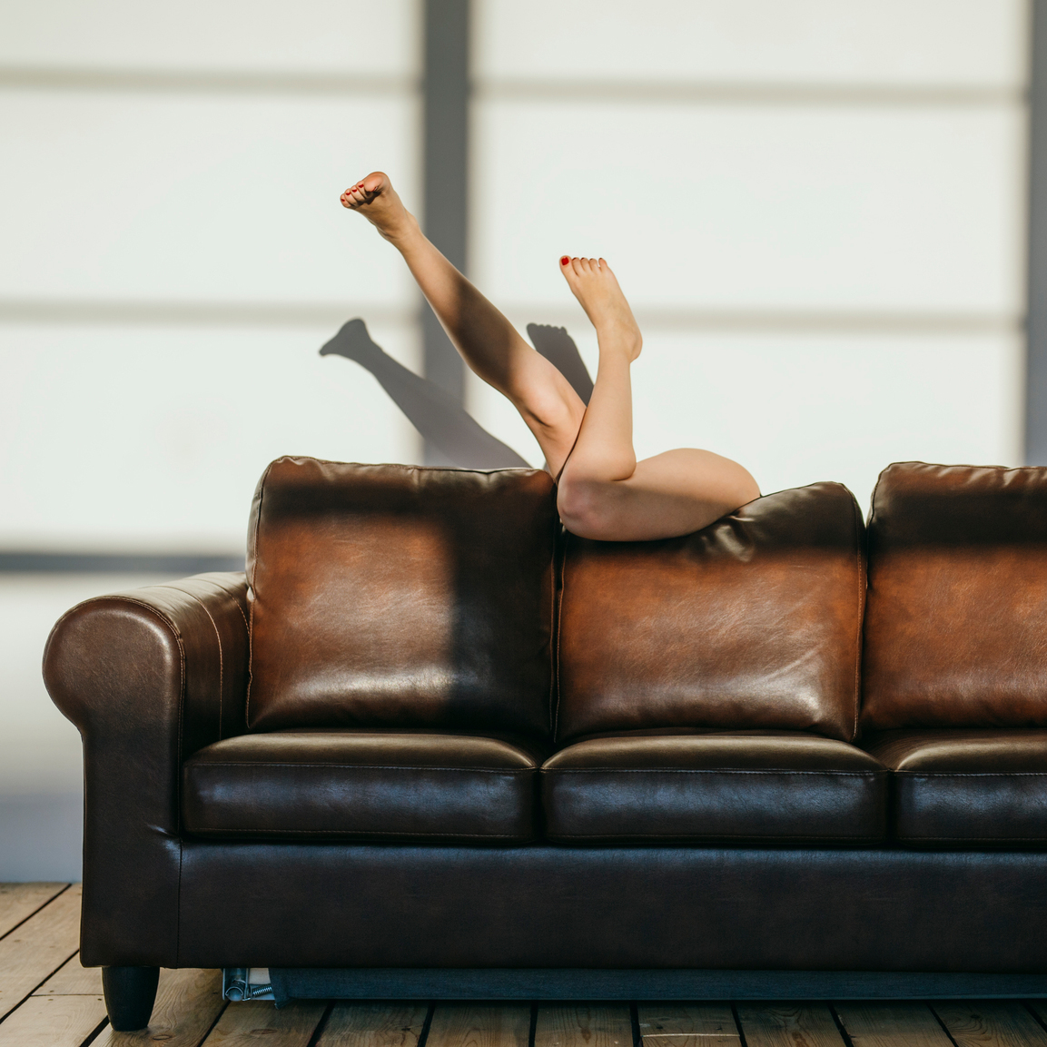 Sexy woman at home under direct sunlight slippage behind brown leather couch