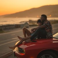 single dad on a date sitting with woman on hood of red car looking at the sunset over the ocean