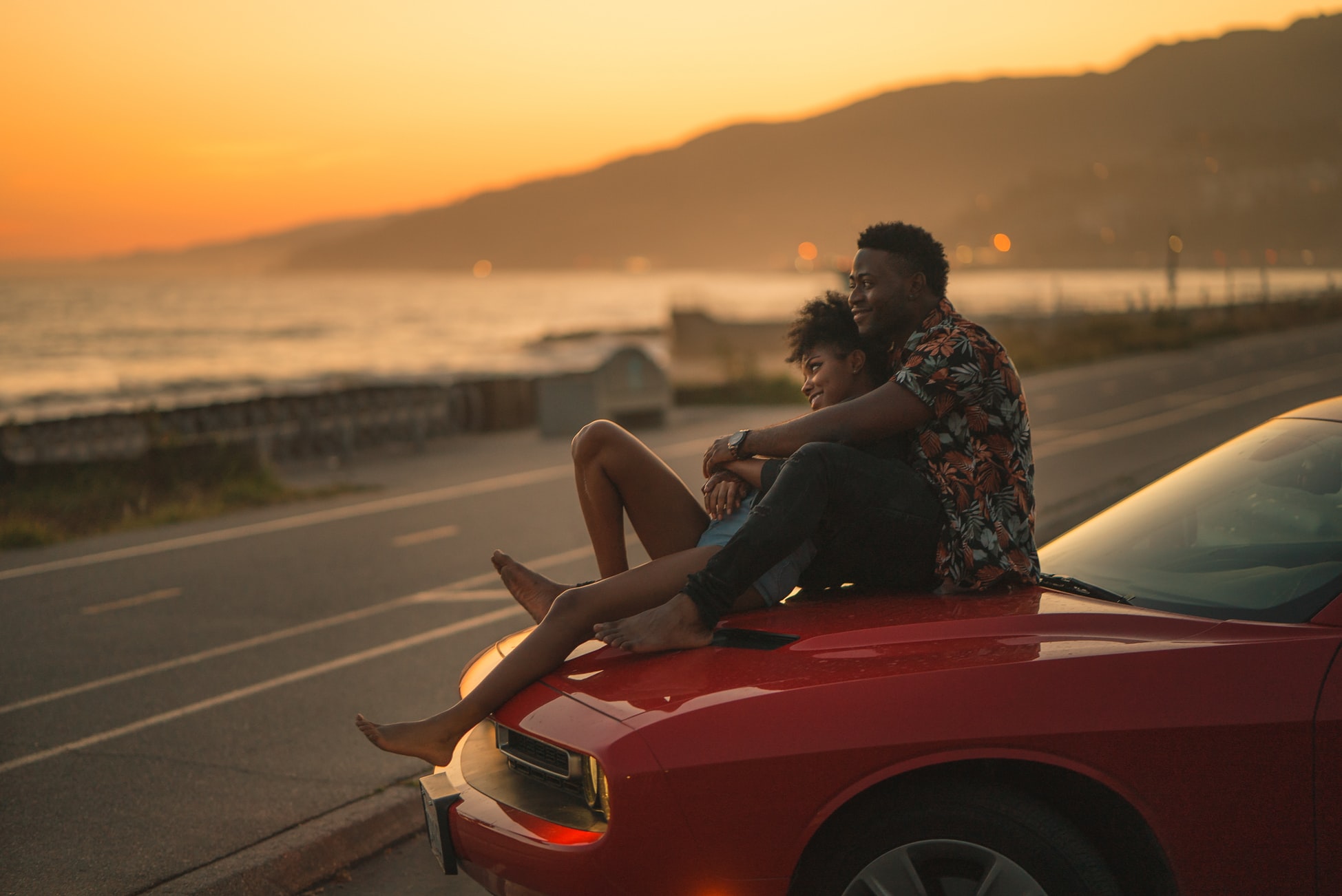 single dad on a date sitting with woman on hood of red car looking at the sunset over the ocean