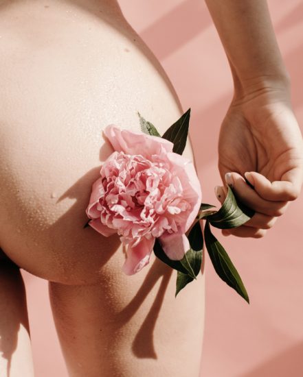 Crop nude female touching buttock with delicate flower against pink background