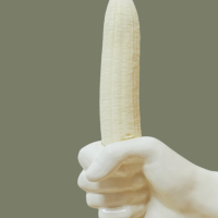 Ceramic hand holding an unpeeled banana against a dark green background.