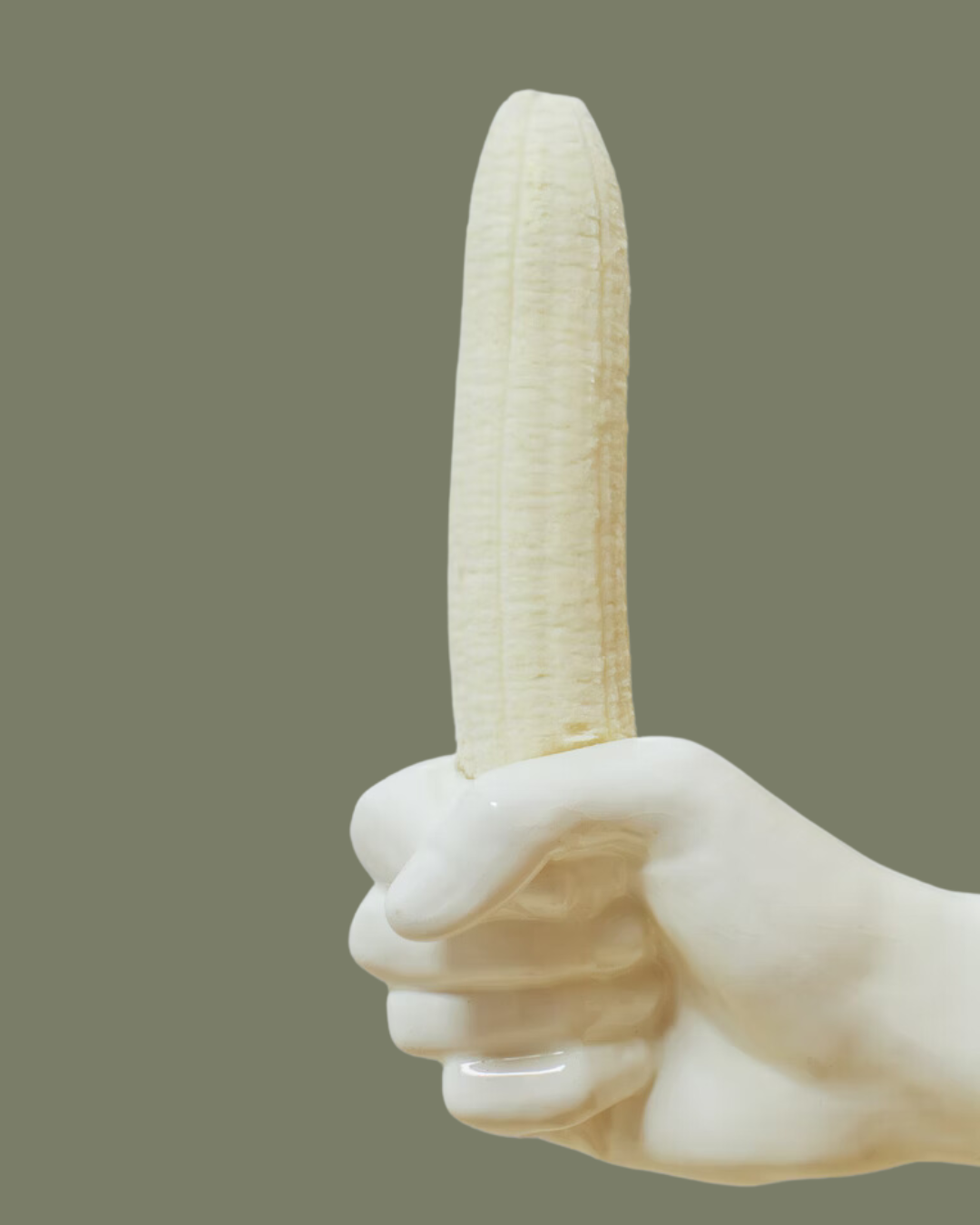 Ceramic hand holding an unpeeled banana against a dark green background.