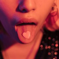 Unrecognizable sensual woman sticking her tongue out with a pink heart. Love concept, orange light.