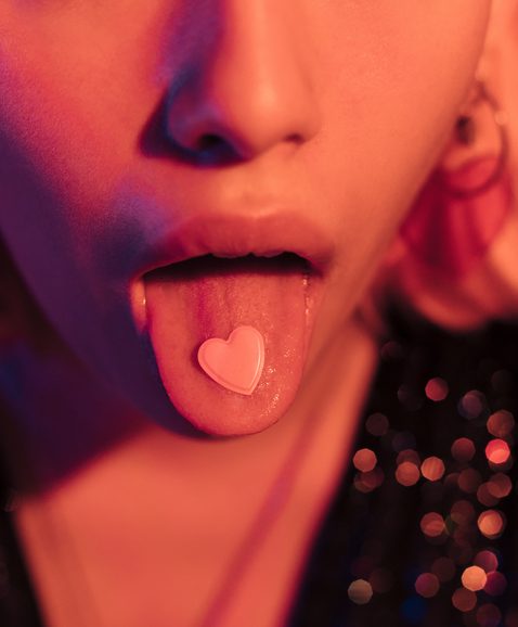 Unrecognizable sensual woman sticking her tongue out with a pink heart. Love concept, orange light.