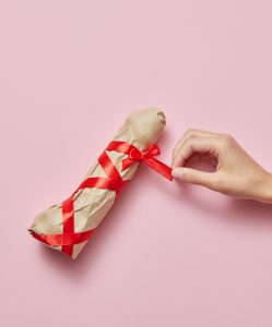Woman's hand unties red silk ribbon on a sex present vibrator wrapped of craft paper as a holiday present on a pastel pink background, copy space. Top view. Gifts and presents concepts.
