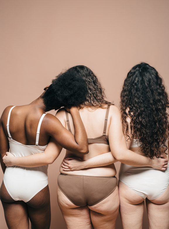 Three mixed races women with their backs turned embracing each other. They wear underwear which show their normal and beautiful bodies.
