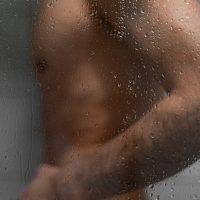 Naked body of anonymous man washing behind wet glass panel with drops of water in shower room.