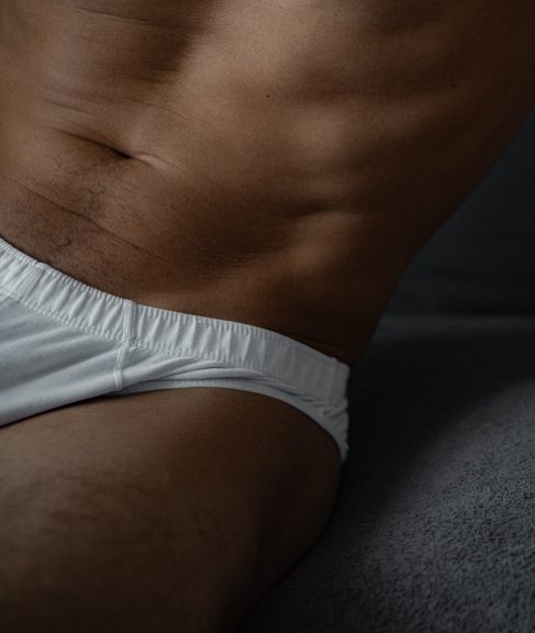 A naked athletic man in white underwear sits freely and confidently on the couch.