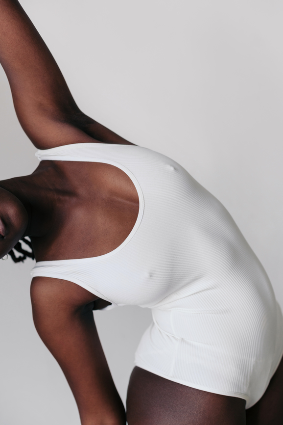 Person in a white ribbed bodysuit stretching gracefully against a neutral background.