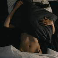 Intimate, dark overhead shot of a person hiding under a large dark pillow in bed, showing bare midriff and hands, symbolizing introspection or hidden feelings.