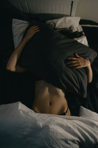 Intimate, dark overhead shot of a person hiding under a large dark pillow in bed, showing bare midriff and hands, symbolizing introspection or hidden feelings.