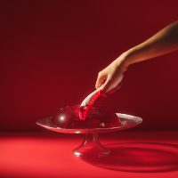 Hand holding a glowing LELO Mona Spectra red light G-spot vibrator dipping into a red jelly dessert