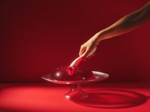Hand holding a glowing LELO Mona Spectra red light G-spot vibrator dipping into a red jelly dessert