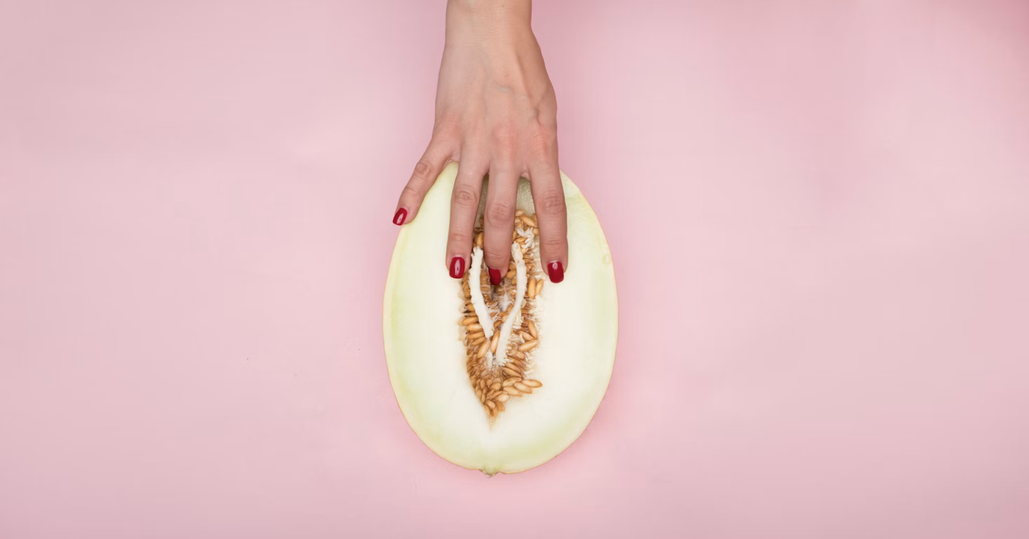 A hand with red manicured nails gently touching the center of a halved melon on a pink background, artistically representing clitoris anatomy and vulva self-touch.
