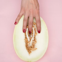 A hand with red manicured nails gently touching the center of a halved melon on a pink background, artistically representing clitoris anatomy and vulva self-touch.