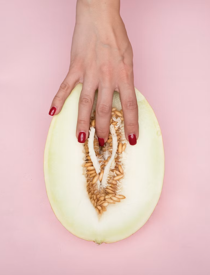 A hand with red manicured nails gently touching the center of a halved melon on a pink background, artistically representing clitoris anatomy and vulva self-touch.